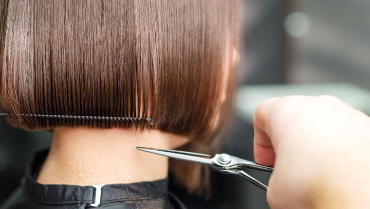 A close-up of a hair stylist’s hand holding scissors as they trim a woman’s hair. 