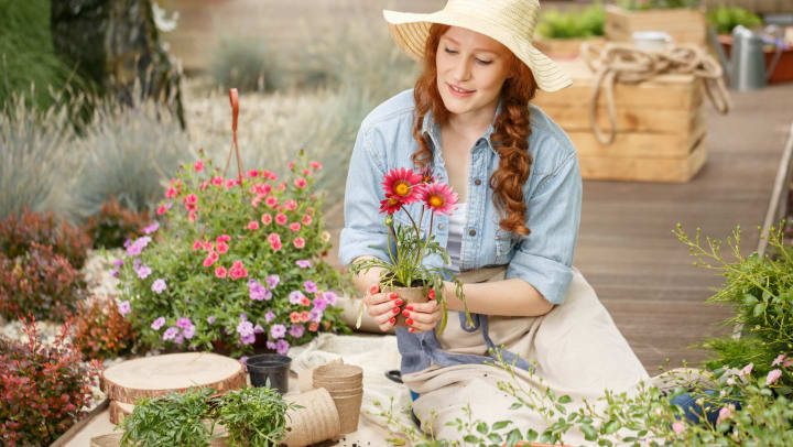 A young woman sitting on a deck holds a pot with a flower in it, while surrounded by a variety of other potted plants and flowers. 