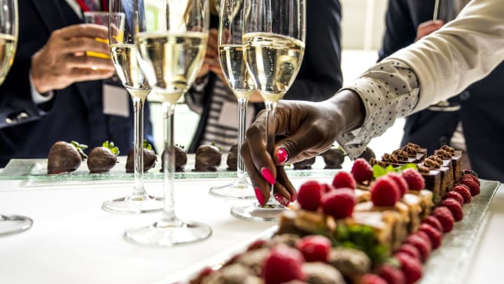 Woman reaching for a champagne glass from a table full of drinks and desserts surrounded by people.