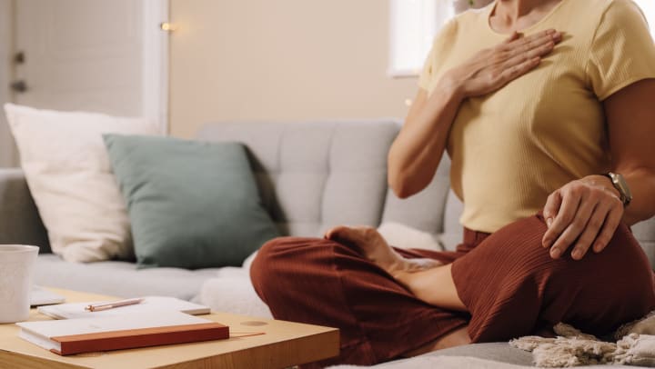 Woman holding a hand to her chest while sitting in a meditative pose on the couch. 