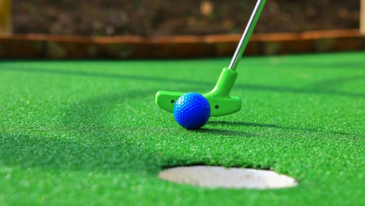 Closeup of green putter hitting blue golf ball toward hole on mini golf course