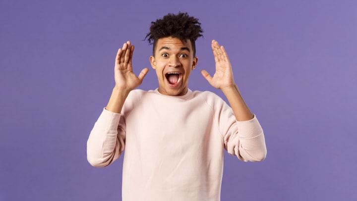 Young man in a pink sweater with his hands up and face making a surprised look.