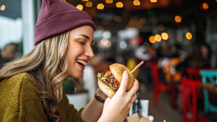 Young woman wearing a purple beanie and olive green sweater smiling and holding a burger in a casual restaurant