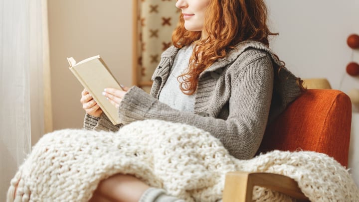 Woman sitting on a chair, with a blanket draped across her lap, looking at an open book in her hands. 
