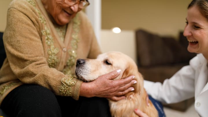 A smiling senior woman and her caregiver pet a therapy dog.