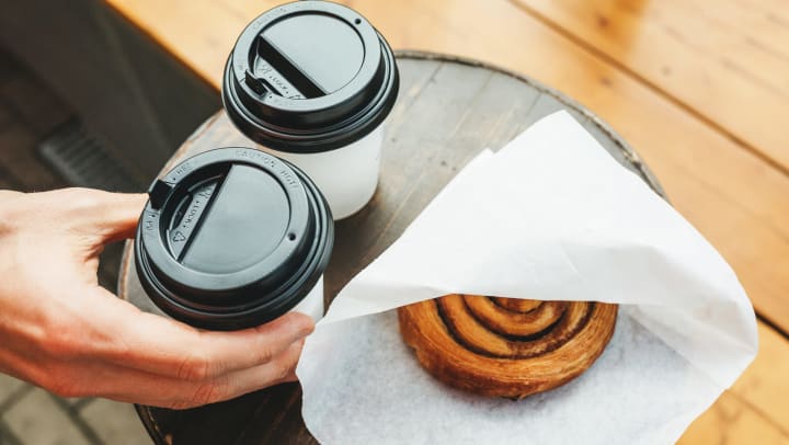 A hand holding a to-go drink cup with black lids on a table next to another to-go cup and a bun in a paper bag.