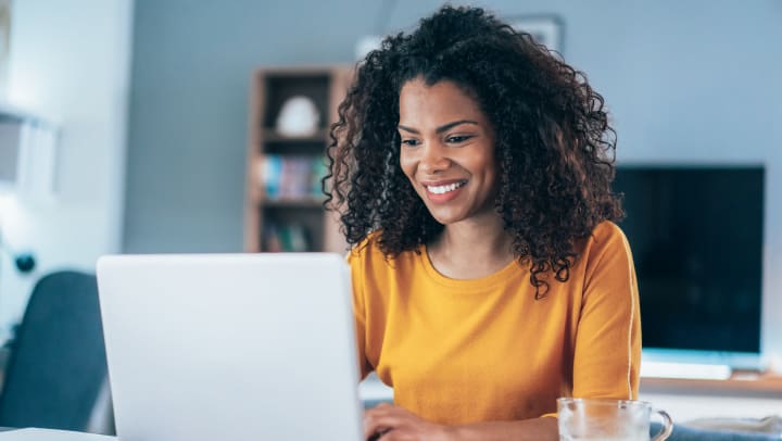 A smiling young woman works on her laptop from a desk in her living room, with a cup of coffee next to her. . 