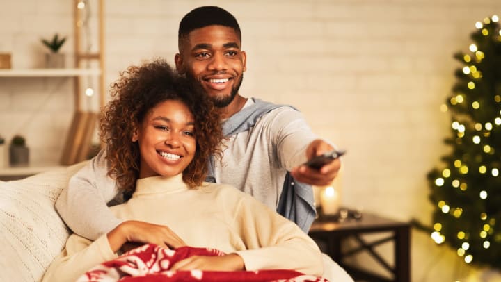 A smiling couple cuddle on the couch under a blanket, as he uses the TV remote. 