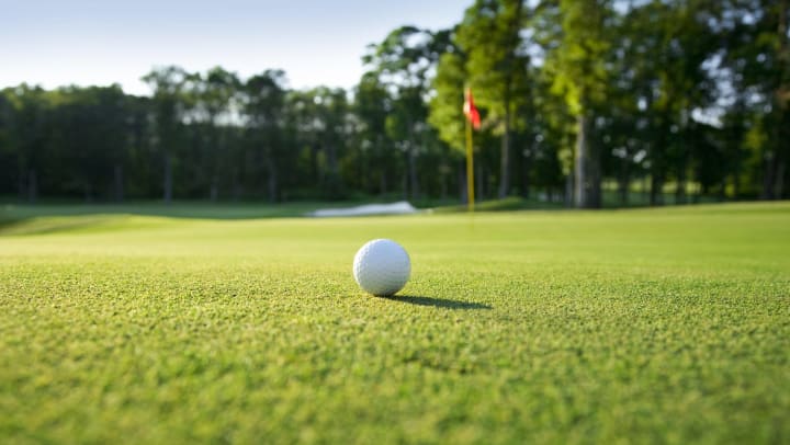 Close up of golf-ball on green with flag and trees in background