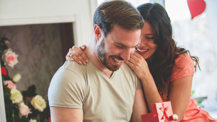 A man opening a small red box. A woman has her arm around him, smiling. 