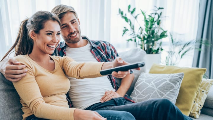 A man and a woman on a couch. The man has his arm around the woman’s shoulder. The woman is holding a TV remote. 