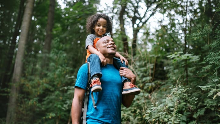 A smiling father carries son on his shoulders during a hike through a forest trail.