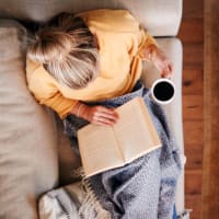 A woman holding a cup of coffee while lounging on her couch reading a book at Astoria in Mobile, Alabama