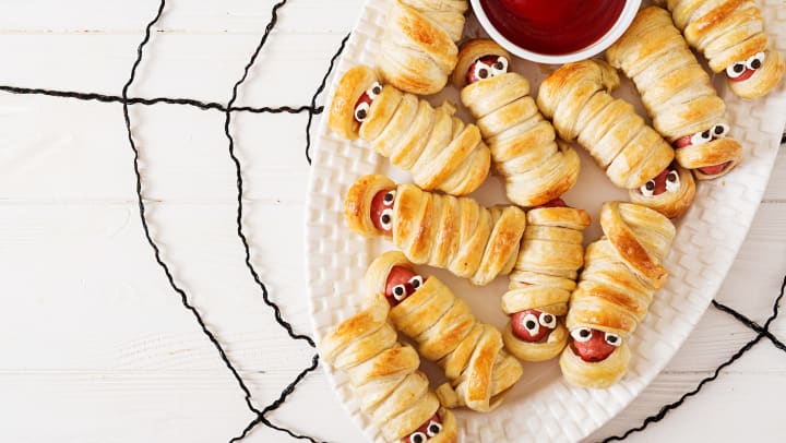 Hot dogs wrapped in pastry puffs to look like mummies sitting on a white plate with a bowl of ketchup.