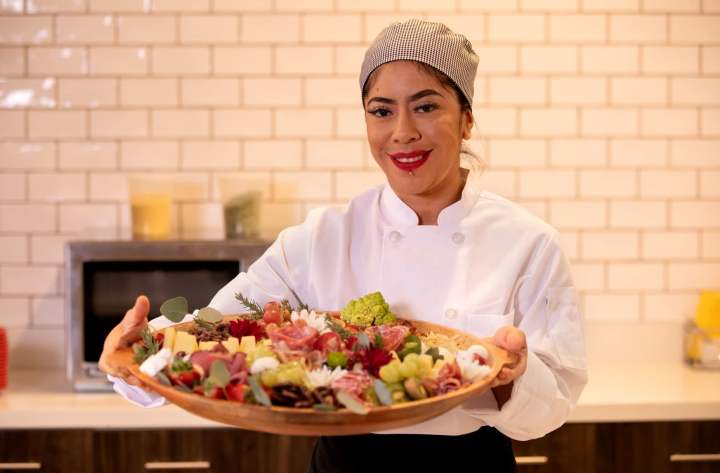 Smiling Clearwater Living chef in a white coat presenting a colorful charcuterie platter filled with fresh fruit, vegetables, and cured meats in a modern kitchen.