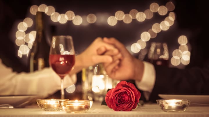 A table with a rose and glass of wine in the foreground and a man’s hand gently holding a woman’s hand. 
