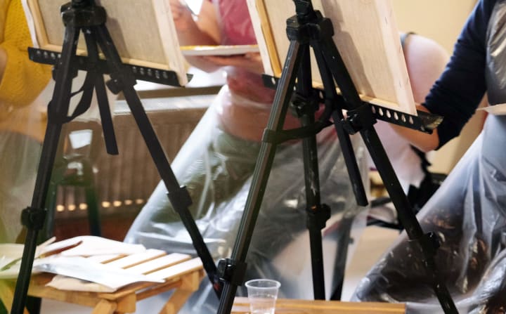 Three art students sitting behind their black easels with bamboo fold out TV trays holding miscellaneous art supplies