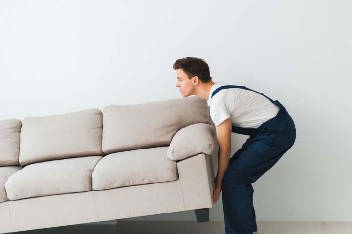 A man in overalls lifts one end of a sofa while moving by himself 