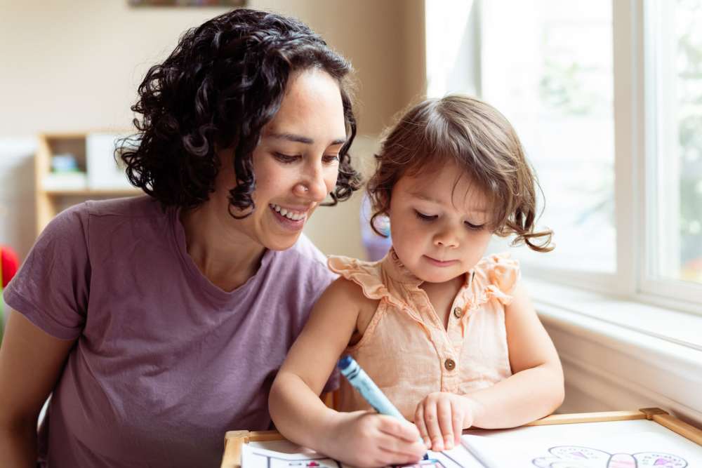 Woman and toddler coloring together in a sunlit living room with large windows at Union Place Apartments in Lafayette, Indiana
