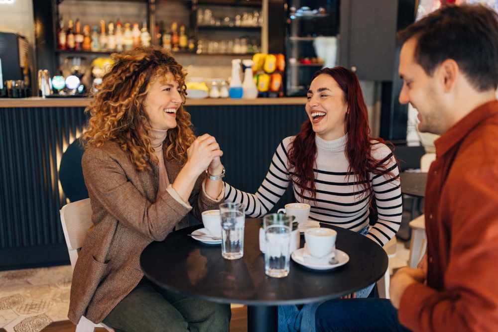 Three friends laughing together at a table with coffee cups near The Waters at Conway in Gonzales, Louisiana