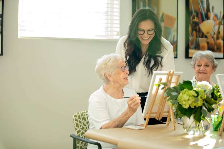 Resident paints at a tabletop easel as a Clearwater team member offers guidance in Clearwater Agritopia’s light filled art studio; another resident looks on.