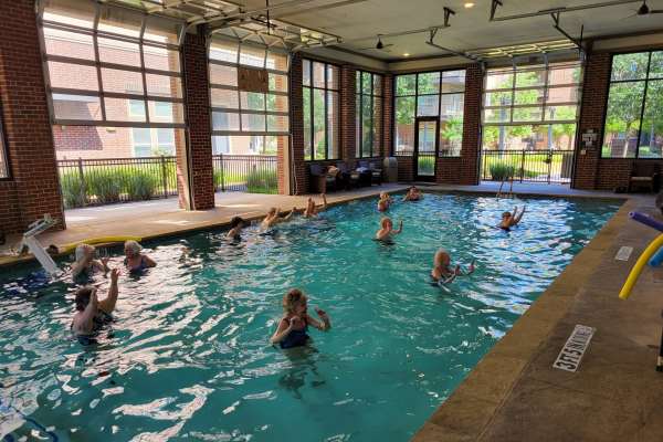 Seniors participating in a water aerobics class in a community pool, with lush greenery and trees visible outside through large windows.