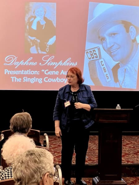 Daphne Simpkins with red hair and black pant suit giving lecture at podium to residents