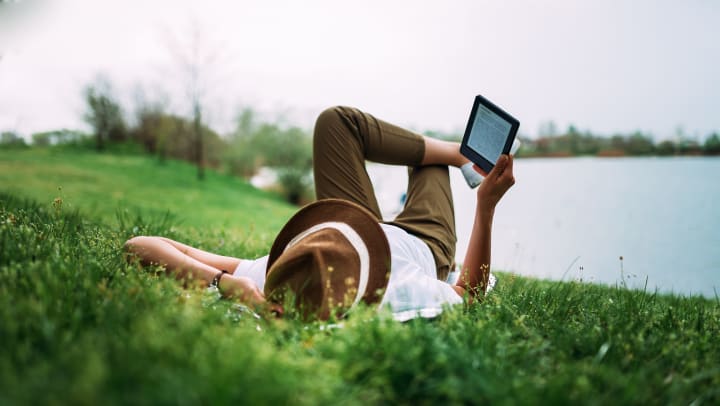 Person wearing a hat while lying in the grass and holding an e-reader. 