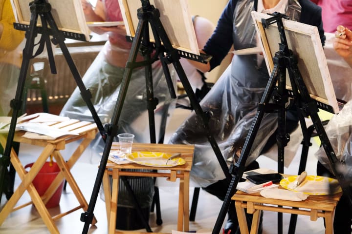 Three art students sitting behind their black easels with bamboo fold out TV trays holding miscellaneous art supplies
