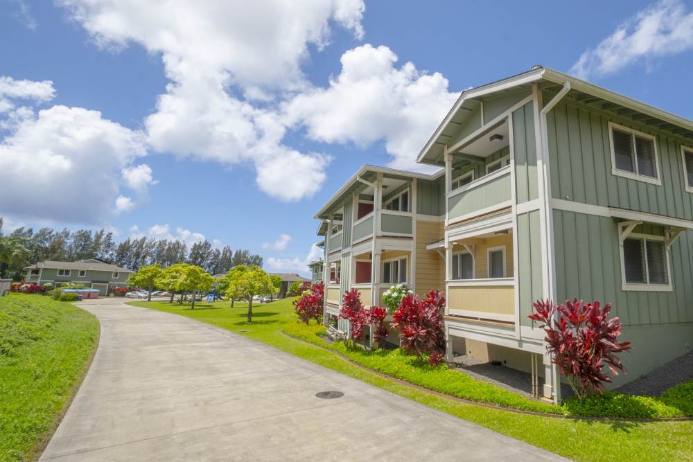 Two-story houses with green and beige siding surrounded by vibrant red shrubs and lush grass under a bright blue sky with scattered clouds at Pa'anau Village II in Koloa, Hawaii