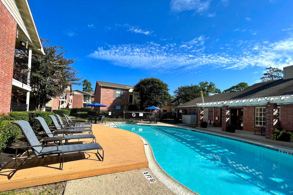 Sparkling Swimming Pool Surrounded By Sun Loungers, Green Shrubs, And Brick Apartment Buildings Under A Clear Blue Sky at The Abbey at Montgomery Park in Conroe, Texas