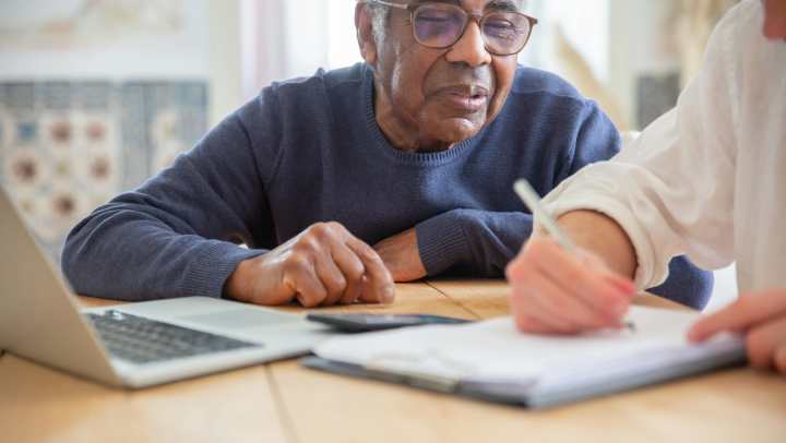 Elderly man on a laptop while another man writes with pen and paper