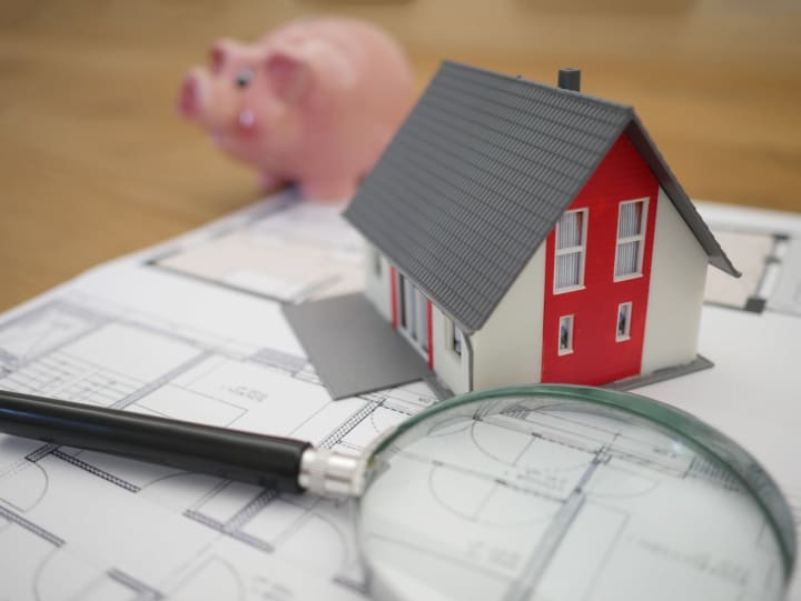 a modular toy sized home sits on top of paperwork with a magnifying glass and piggy bank in the background