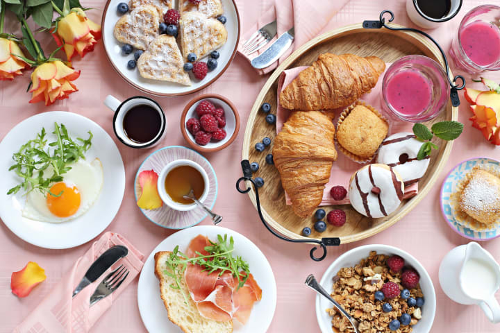 A table spread of croissants, donuts, waffles, mixed berries, eggs, coffee, and other brunch entrees on a pink tablecloth