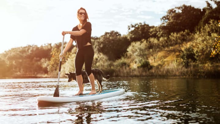 Young woman in black athletic clothes holding a paddle and standing on a stand-up paddleboard with her dog on a sunlit river