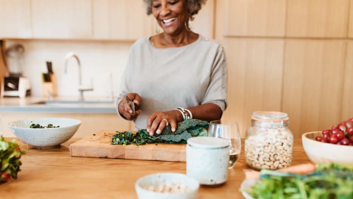 Elderly woman in a kitchen smiling while holding a knife over some chopped kale on a cutting board.
