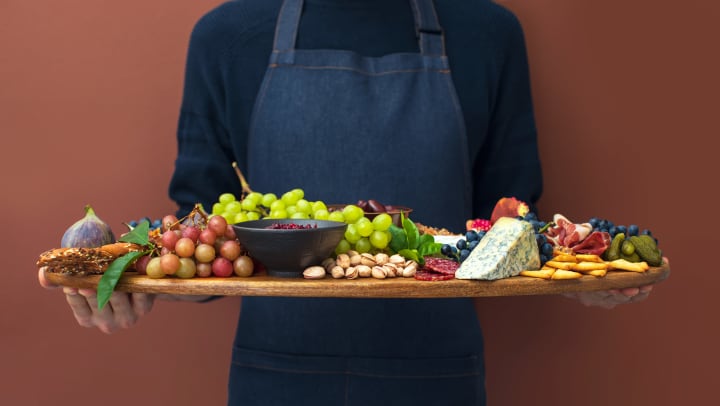 Man in an apron holding a charcuterie board with grapes, cheese, crackers, meat, and pickled food items