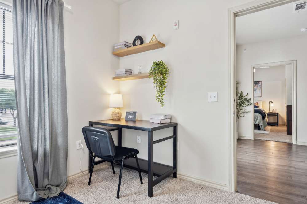 Study room with black desk, shelves, lamp, and books; open doorway reveals bedroom with hardwood flooring at Midtown 905 in Denton, Texas