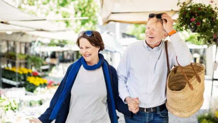 A smiling, older couple walking through a flower market on the street while holding hands. 