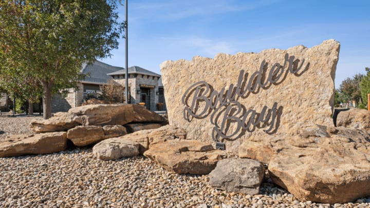  A photo of the entrance sign and clubhouse of the Boulder Bay Apartments in Amarillo, TX, under a bright blue sky with white clouds. In the foreground right, a large, light-colored stone monument sign has the community name "Boulder Bay" in cursive, dimensional lettering. The foreground landscaping consists of extensive brown and gray gravel. To the left, a flagpole holds the US flag and other flags. In the background, the single-story clubhouse building has a gray shingled roof, a light brown stone and brick exterior, and a prominent entrance canopy.