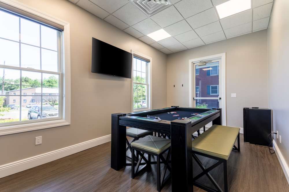 Indoor game room with a poker table, chairs, wall‑mounted television, large windows, and wood‑look flooring at Center Grove Village in Randolph, New Jersey.