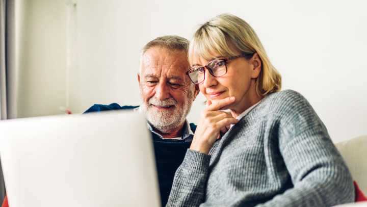 Two smiling seniors looking at a computer together, exploring online resources in an Independent Living community.