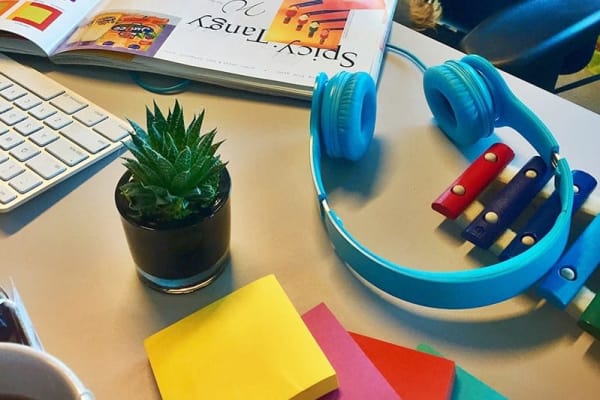 Small green desk plant, multi-colored sticky notes, rainbow colored children's toy xylophone, blue headphones, open book, and computer keyboard scattered on desk.