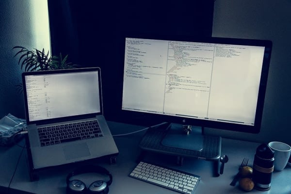 Notebook computer and display, wireless headphones and keyboard, fork, spoon, 2 tangerines, thermos, and a coffee cup sitting at a desktop workstation.