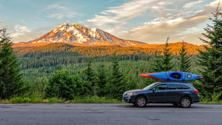 A sport utility vehicle carrying kayaks travels down a road with a forest and mountain in the background