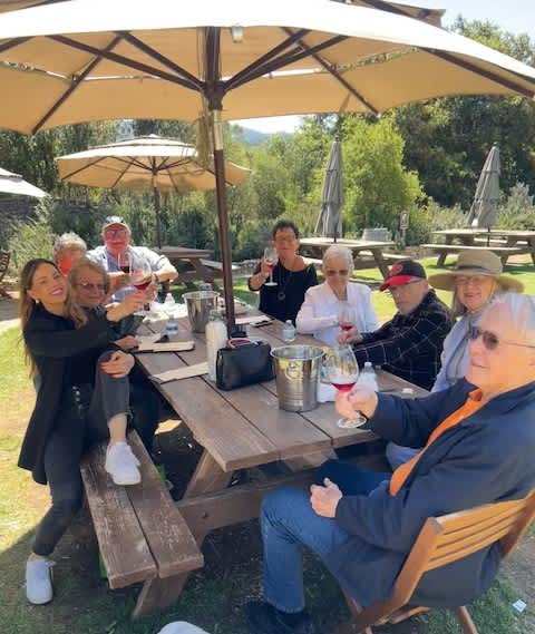 Clearwater at Riverpark residents sit at a shaded picnic table under umbrellas, raising tasting glasses during an outdoor wine outing with notes and an ice bucket on the table.