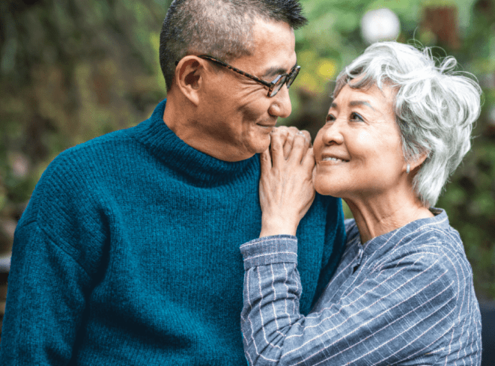 Older Asian couple smiling warmly at each other, standing close outdoors in a peaceful, natural setting.
