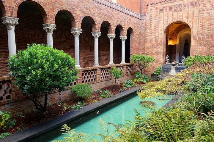 A view of the courtyard inside of the Jordan Schnitzer Museum of Art. An all bring building surrounds a small courtyard filled with plants and a small crystalline blue rectangular body of water sits in the middle of the foliage. 