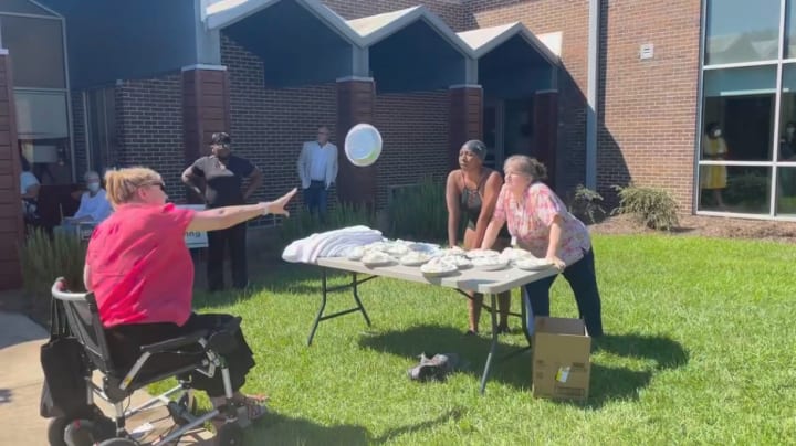 Woman in wheelchair outside hurls carnival pie toward two staff members ready to accept the blow