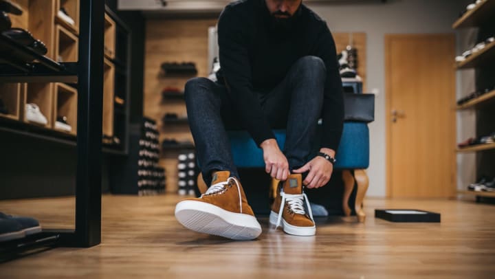 young man tries on suede, high-top sneakers while sitting down at a trendy shoe store | footwear stores in Keller